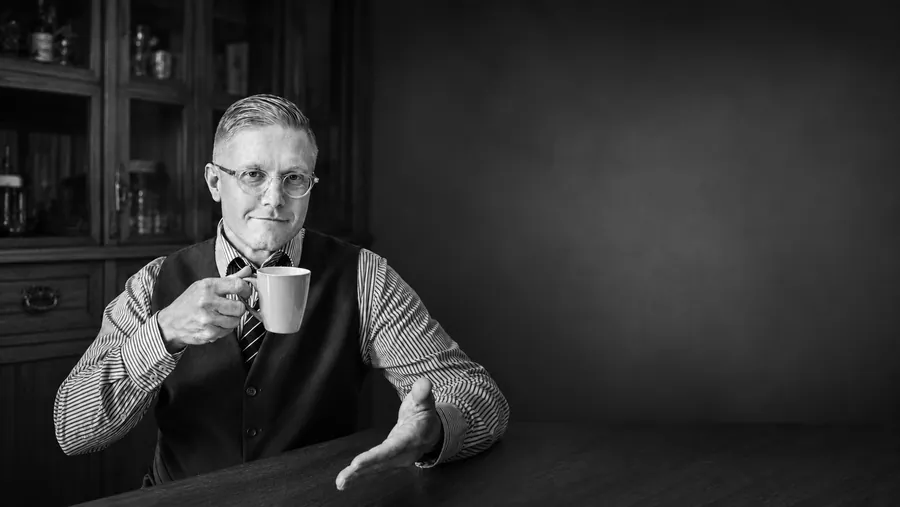 Dennis Holewa am Tisch mit einer Kaffeetasse, die andere Hand einladend geöffnet